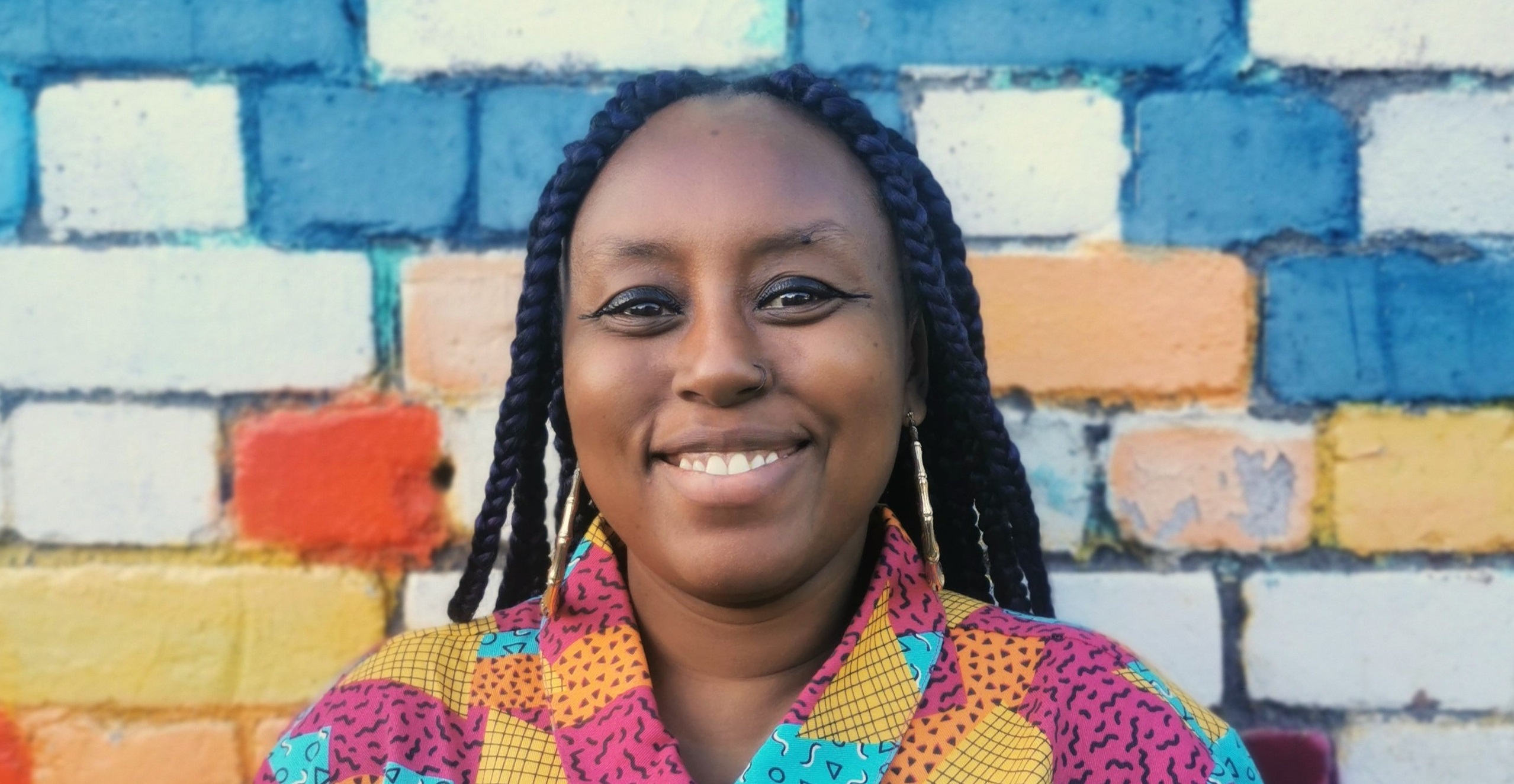 An image of graphic artist Nadina Ali, who is a black female with long hair in dreadlocks smiling in front of a colourful background. She is wearing a colourful patchwork shirt.